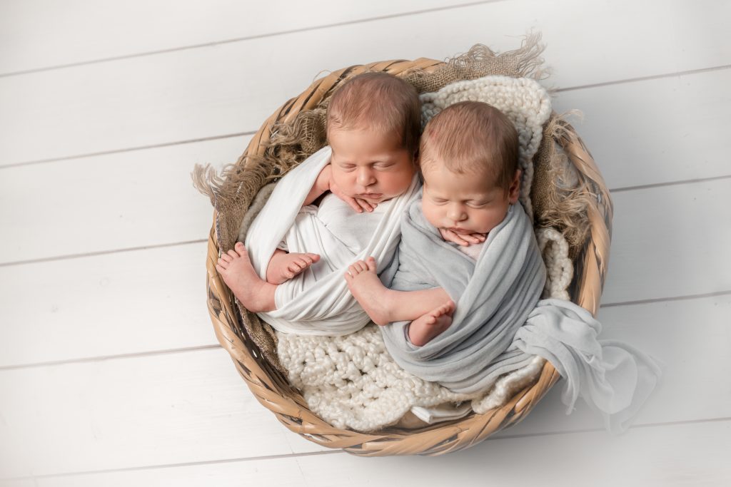 Gémeos recém-nascidos a dormir num cesto de vime, envoltos em panos branco e cinzento, numa sessão newborn Pose in Studio em Leça da Palmeira, Porto