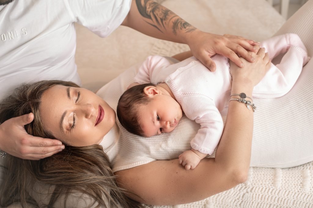 Mãe deitada de olhos fechados com a bebé recém-nascida vestida de rosa ao peito e o pai a acariciar-lhes a cabeça, sessão newborn Natural / Lifestyle no Porto